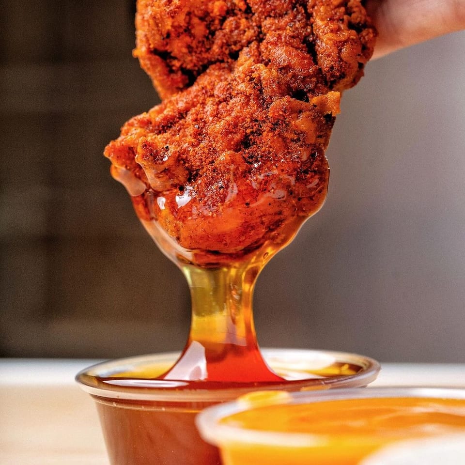 Crispy, heavily seasoned fried chicken tender held above cups of dipping sauces.