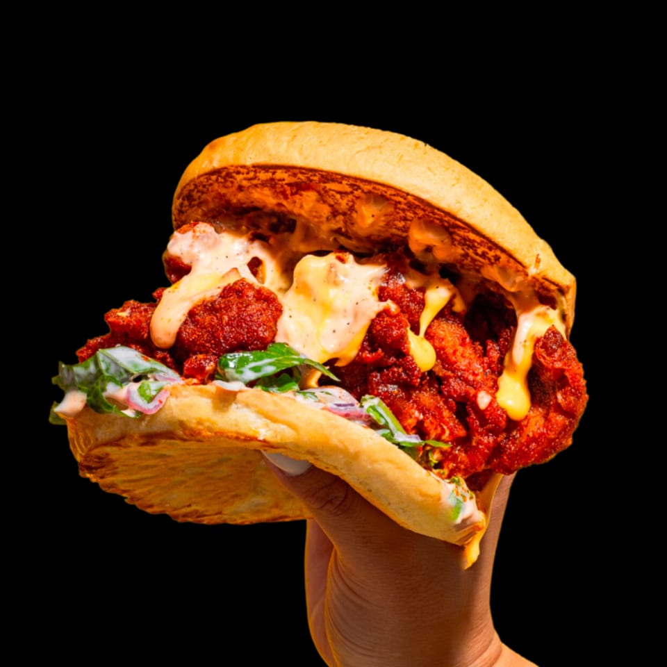 Hand holding a loaded fried chicken sandwich with creamy sauces, greens, and pickled onions against a black background.