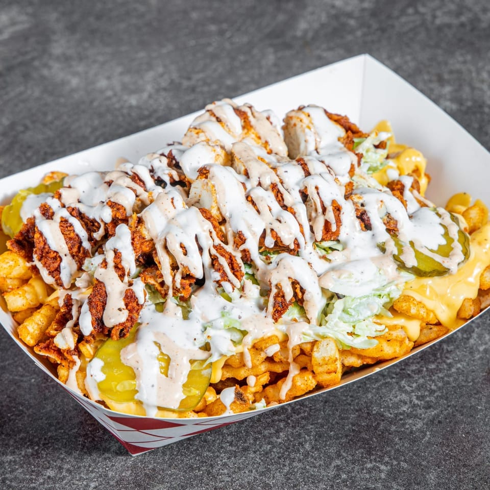 Loaded curly fries topped with seasoned chicken, lettuce, pickles, cheese sauce, and creamy white dressing in a paper tray.