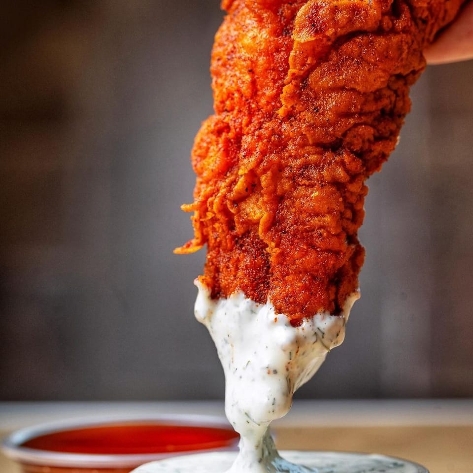 Crispy spicy fried chicken tender dipped in creamy herb sauce, with red and yellow dipping sauces nearby.