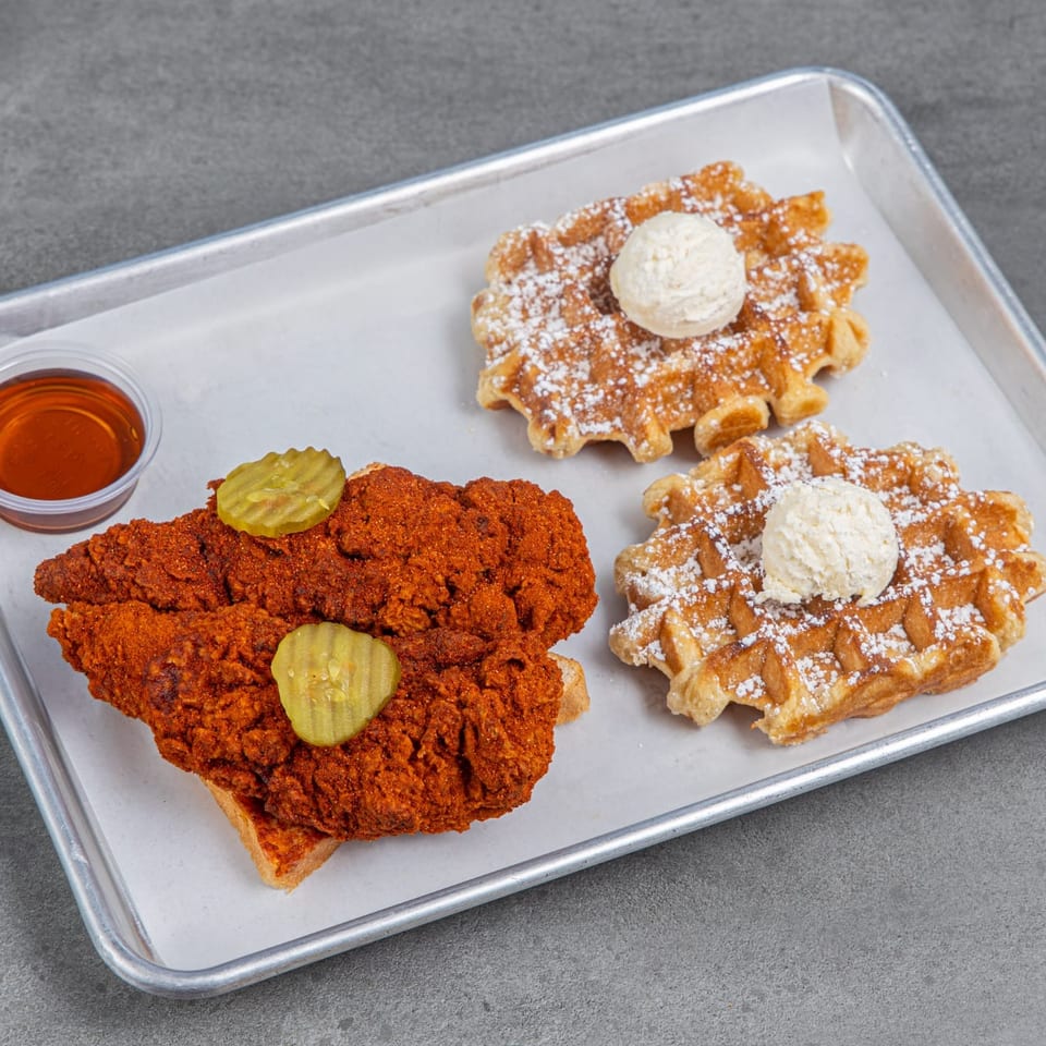 Tray of spicy fried chicken and powdered-sugar waffles topped with vanilla ice cream, pickles, and syrup.