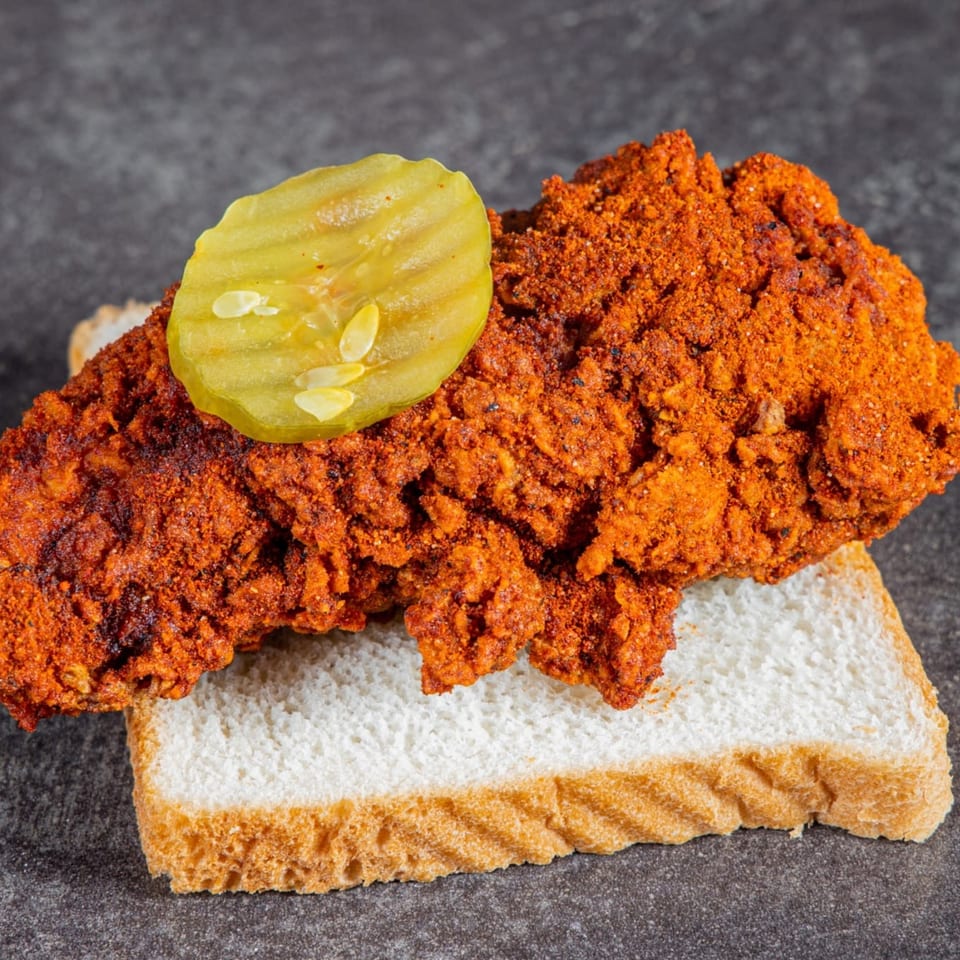 Spicy crispy fried chicken piled on white bread and topped with a pickle slice.