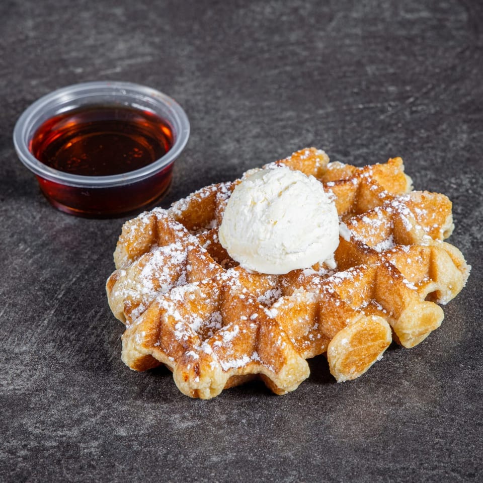 Golden waffles dusted with powdered sugar, topped with whipped cream, and served with a cup of syrup.