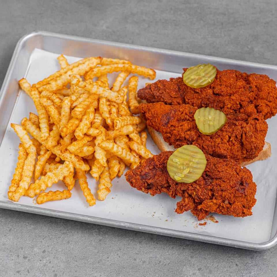 Spicy fried chicken tenders with pickle slices and seasoned crinkle-cut fries served on a paper-lined metal tray.